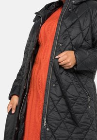 Mujer vistiendo un abrigo negro acolchado parcialmente abierto, revelando un vestido de punto de cable naranja debajo, sosteniendo la solapa del abrigo con una mano.