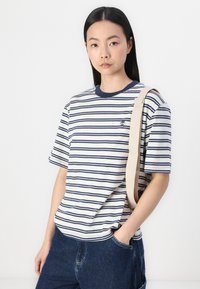Striped short-sleeve T-shirt in cream and navy blue, featuring a crew neck and a small logo on the chest. Paired with dark denim pants.