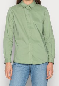 En person står iförd en ljusgrön skjorta med knappar och blå jeans, med avslappnade händer vid sidorna mot en enkel vit bakgrund.