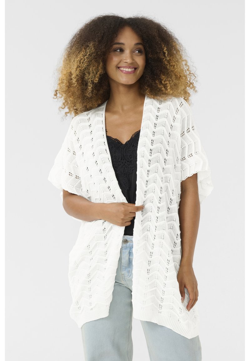 Vit öppen stickad cardigan med en chevronmönster, korta ärmar och en lös passform. Bärs över en svart topp med ljusblå jeans.