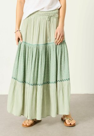 Frau trägt einen langen, gestuften grünen Rock mit gestreiften und floralen Mustern, kombiniert mit braunen Sandalen und einem weißen Oberteil.