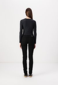 Frau mit langen, glatten Haaren, die ein eng anliegendes schwarzes Langarmoberteil, schwarze enge Jeans und schwarze Schuhe trägt und vor einer weißen Wand steht.