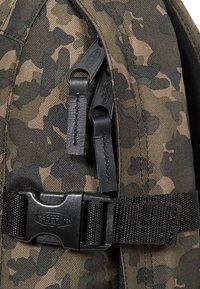 Plecak w wzór kamuflażowy, wykonany z trwałego materiału, z czarnymi suwakami i okuciami, regulowanymi szelkami oraz zapięciem na plastikową klamrę.