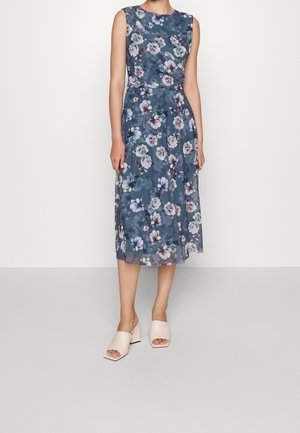 Niebieska sukienka midi bez rękawów z różowo-białym kwiatowym wzorem, noszona z białymi sandałami na obcasie typu blok z odkrytymi palcami, modelka stoi na jednolitym tle.