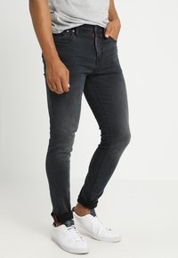 Jeans de denim negro ajustados con un leve desgaste, diseño de cinco bolsillos y costuras rojas, combinados con zapatillas blancas y una camisa gris.