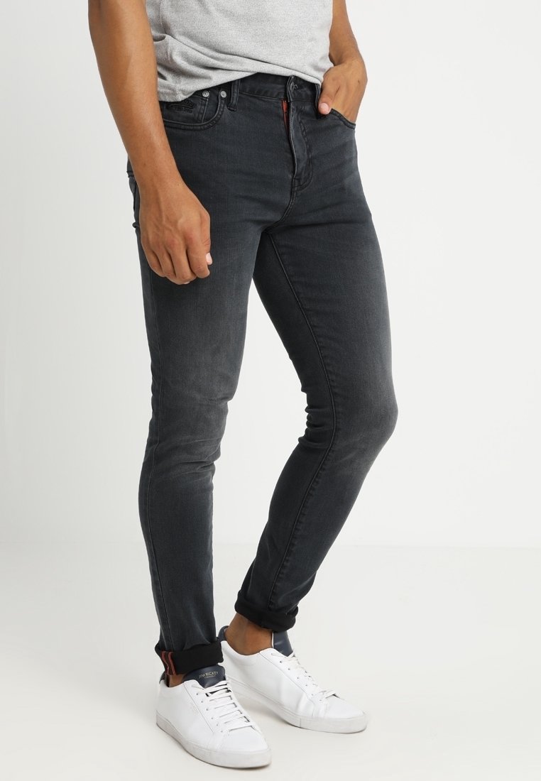 Jeans de denim negro ajustados con un leve desgaste, diseño de cinco bolsillos y costuras rojas, combinados con zapatillas blancas y una camisa gris.