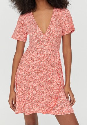 Femme portant une robe portefeuille corail à manches courtes et col en V, imprimée de petites fleurs blanches, debout avec les mains détendues le long du corps.