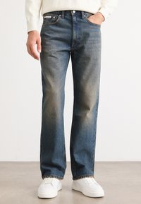 Jeans de mezclilla azules con un diseño de pierna recta, ligero desgaste y un estilo clásico de cinco bolsillos. Combinados con zapatillas blancas.