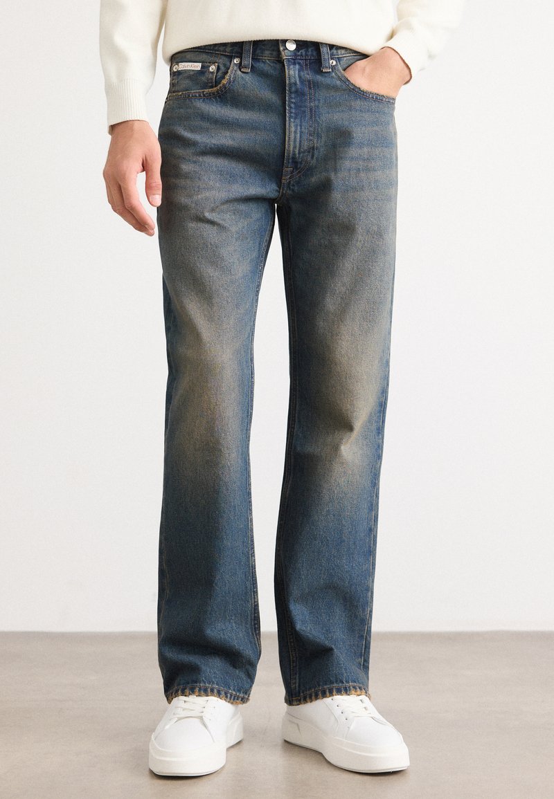 Jeans de mezclilla azules con un diseño de pierna recta, ligero desgaste y un estilo clásico de cinco bolsillos. Combinados con zapatillas blancas.