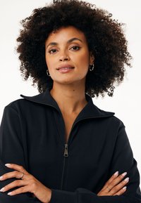 Chaqueta negra con cremallera y cuello alto, fabricada con un material suave y con una cremallera lisa. El modelo muestra los brazos cruzados y un fondo neutro.