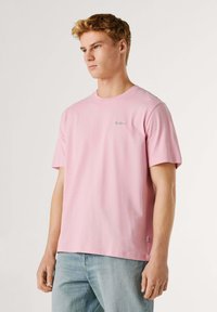 Jeune homme aux cheveux blonds courts portant un t-shirt rose uni à manches courtes et un jean bleu clair, regardant sur le côté sur un fond blanc.