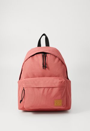 Pink fabric backpack with a rounded top, adjustable black straps, front pocket, and a tan logo tag. Smooth texture and minimalistic design.