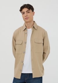 Camicia button-up di un marrone chiaro, realizzata in un tessuto morbido, con due tasche sul petto e un colletto classico, abbinata a una maglietta bianca sotto.