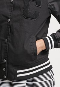 Chaqueta universitaria negra con puños y dobladillo a rayas blancas, gran letra "C" texturizada en el pecho, mano en el bolsillo lateral, combinada con pantalones a rayas negras y blancas.