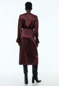 Bordeauxfarbendes Satin-Kleid mit langen Ärmeln, cinchierter Taille und geknöpften Manschetten, kombiniert mit eleganten schwarzen kniehohen Stiefeln. Glatte Textur, knielanger Stil.