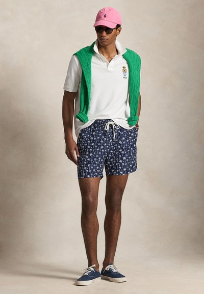 Mann iført rosa caps, solbriller, hvit poloskjorte med bjørnelogo, grønn genser over skuldrene, marineblå shorts med palmebladprint og blå joggesko.