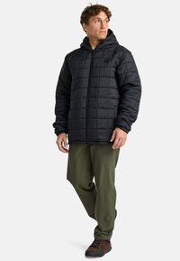 Hombre vestido con una chaqueta con capucha acolchada negra, pantalones verde olivo y zapatos de senderismo marrones, de pie con las piernas cruzadas y mirando de lado.