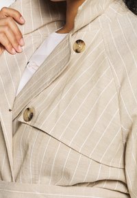 Beige linnen jas met witte strepen, voorzien van een grote kraag en twee ronde knopen. De textuur lijkt lichtgewicht en op maat gemaakt.