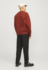 Pull en tricot couleur rouille avec un ourlet côtelé, associé à un pantalon noir et des chaussures noires. Le mannequin porte une casquette claire avec une visière verte.
