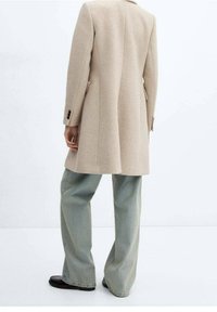Beige uldfrakke med skræddersyet silhuet og spidse revers, kombineret med lyseblå, vide jeans og sorte sko.