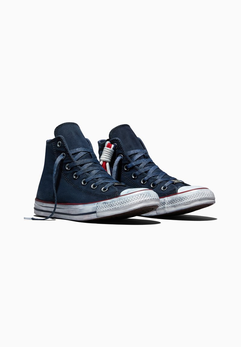 Paar marineblaue High-Top-Sneaker mit weißen Gummisohlen und Schnürsenkeln, rotem Streifendetail an der Sohle, vor weißem Hintergrund.