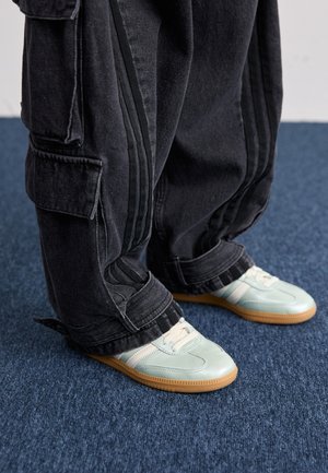 Osoba nosząca jasnozielone sneakersy z białymi paskami i luźne czarne spodnie typu cargo stoi na niebieskim wykładzinowym podłożu.