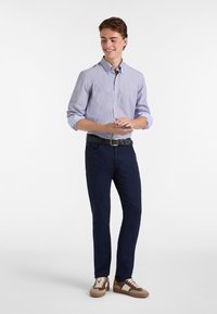 Camicia button-up a righe blu chiaro con maniche arrotolate, chinos blu navy e sneaker multicolori, su uno sfondo bianco puro.