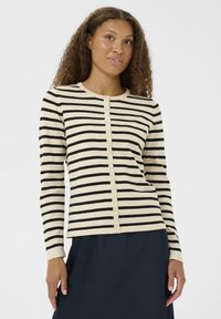 Ženska s kodrastimi lasmi, oblečena v bež in črni horizontalno prugasti kardigan s gumbi in temno krilo, obrnjena rahlo levo.