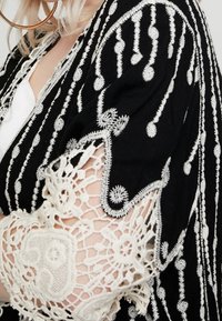 Garbato in tessuto nero con intricati ricami bianchi caratterizzati da motivi circolari e lineari; gli accenti in pizzo sulle maniche aggiungono texture e dettaglio.