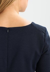 Marineblå klänning med en jämn textur, med en bred båtutringning och randiga axelaccenter. Inkluderar en bakre dragkedjestängning.