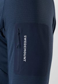 Gros plan sur une manche bleu marine avec une poche zippée et le mot « SWEDEMOUNT » imprimé verticalement en lettres blanches.