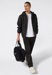 Czarna pikowana kurtka z kapturem, biała koszulka, czarne spodnie i jasne sneakersy. Nosi czarną torbę podróżną z logo.