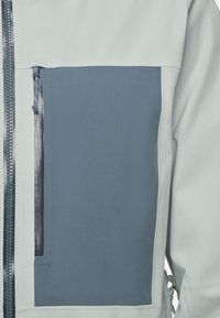 Veste gris clair avec des panneaux bleu marine, tissu texturé, poche zippée et cordons de serrage réglables à la taille.