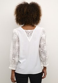 Weißes Blusenshirt mit Spitzenpuffärmeln und V-Ausschnitt hinten. Die Spitze zeigt florale Muster, und der Stoff hat eine glatte Textur.