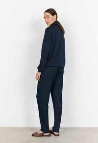 Navyblaues Sweatshirt- und Hosen-Set, aus weichem Stoff, mit hohem Kragen, langen Ärmeln und lockerer Passform. Trägt dazu braune offene Schuhe.