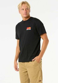 Camiseta negra de manga corta hecha de algodón con un pequeño logo colorido de "RIPCURL" en el pecho. Combinada con pantalones cortos de pana color caqui.