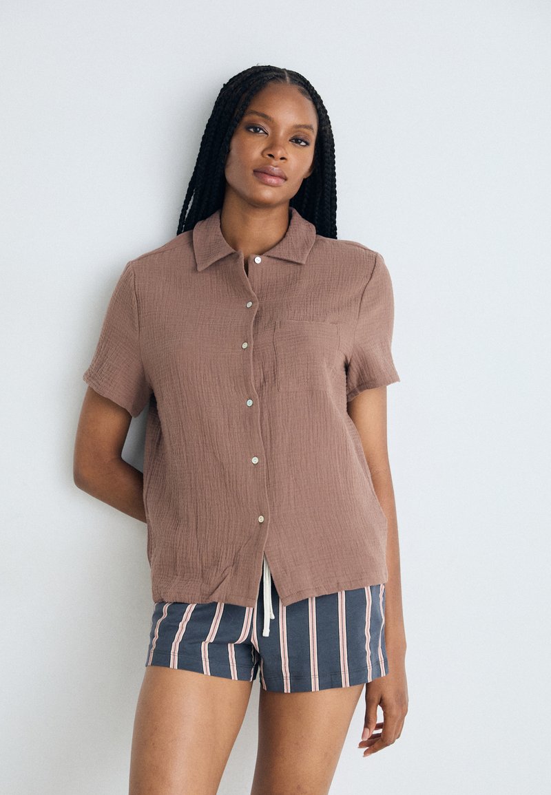 Chemise brune à manches courtes et à boutons en tissu texturé, avec une poche avant et un ourlet légèrement arrondi. Portée avec un short rayé bleu marine.