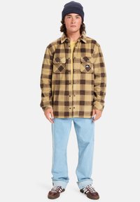 Braun-beige kariertes Hemd mit zwei Brusttaschen, kombiniert mit hellblauen Jeans und braunen Turnschuhen. Getragen mit einer dunklen Mütze.