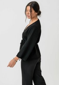 Pulover negru, înfășurat, dintr-un material tricotat moale, cu decolteu în V, cordon în talie și mâneci clopot. Textura este netedă, cu un luciu ușor.