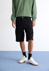 Black cargo shorts with two side pockets, worn with a green long-sleeve shirt and light-colored sneakers. Textured fabric, knee-length.