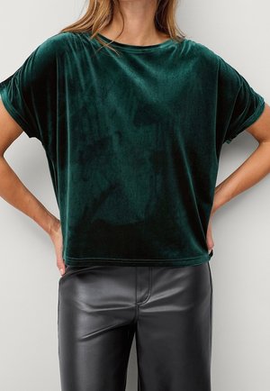 Kvinde iført en løs, mørkegrøn fløjls-t-shirt med korte ærmer, kombineret med sorte læderbukser, stående med hænderne på hofterne.