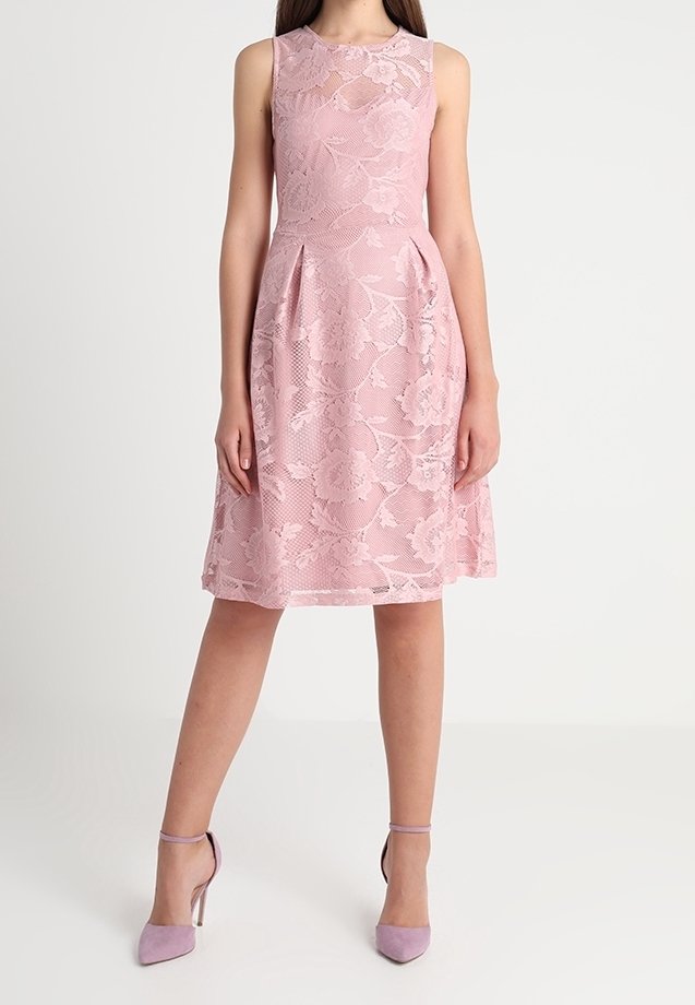 Robe rose en dentelle sans manches longueur genou avec motif floral, portée avec des talons hauts assortis à brides à la cheville, sur une personne debout devant un fond uni.