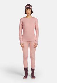 Kobieta stojąca w bladoróżowej bluzce z długim rękawem i legginsach, ciemnofioletowych, luźnych skarpetkach oraz pasującej opasce na głowę z jasnoróżowym napisem.