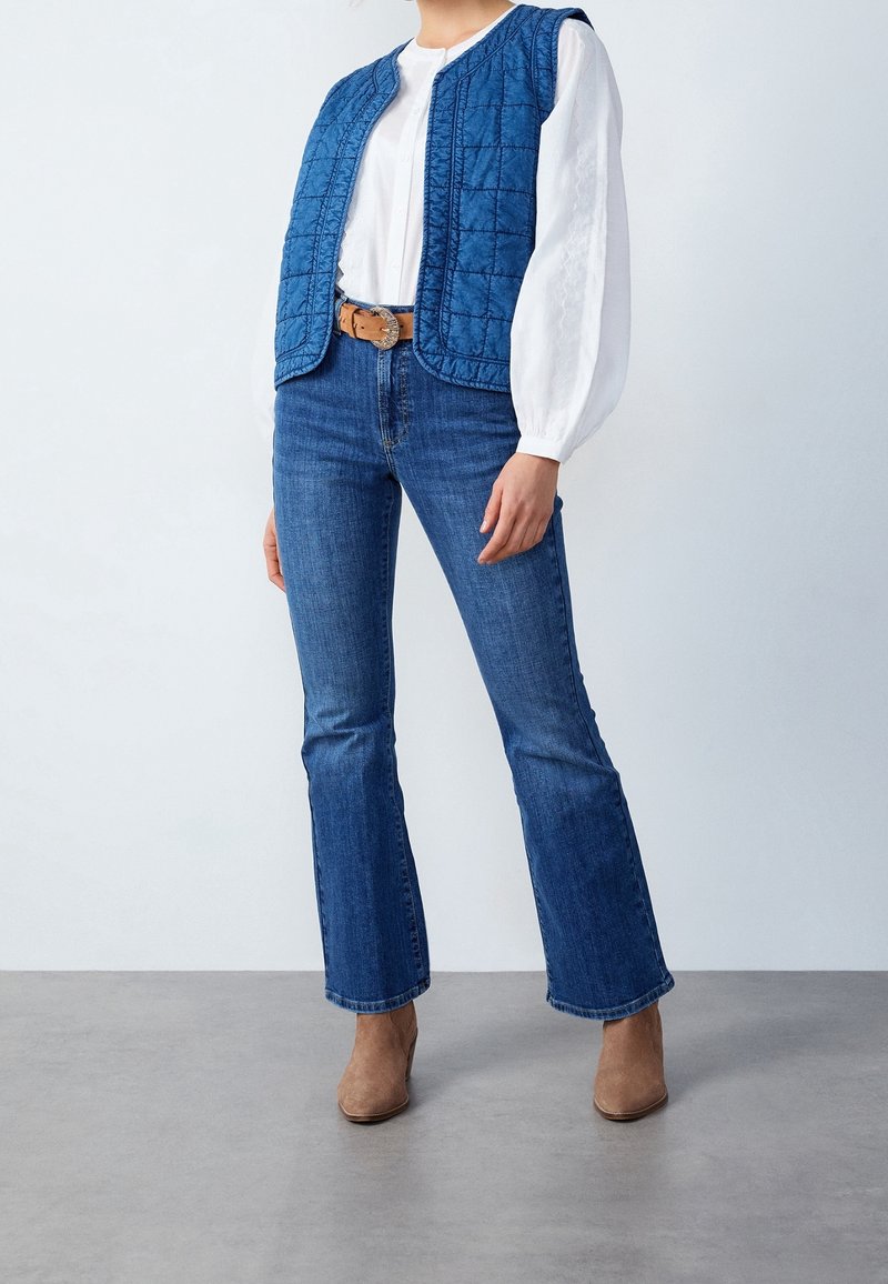 Blå vadderad väst med hög krage, vit långärmad blus, tillsammans med utsvängda blå jeans och bruna anklarstövlar.