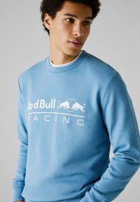 Svijetloplavi dukserica s okruglim izrezom, s bijelim natpisom "Red Bull Racing" i grafikom dvaju bikova na prednjoj strani. Mekan materijal.