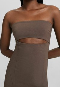 Robe bustier marron foncé avec une découpe à la taille, portée par une personne à la peau foncée et aux cheveux tressés, sur un fond uni.
