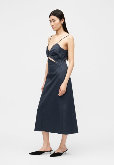 Γυναίκα με μακριά σκούρα μαλλιά που φοράει ένα navy μπλε midi φόρεμα με λεπτές τιράντες, λεπτομέρειες στο στήθος, πλαϊνή έξοδο και μαύρα μυτερά mules με τακούνι kitten heel.
