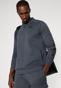 Sudadera gris con cremallera, hecha de un tejido texturizado, que presenta un alto cuello, bolsillos laterales y un logo negro en el pecho.