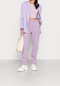 Fialový oversize blazer přes pruhované crop top, doplněný o fialové kalhoty s vysokým pasem a bílé boty s mohutnou podrážkou. Drží krémovou kabelku.