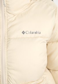 Jachetă puffer bej deschis cu un finisaj texturat, închiderea cu fermoar și logo-ul Columbia pe partea stângă. Dispune de secțiuni cusute pentru izolație.